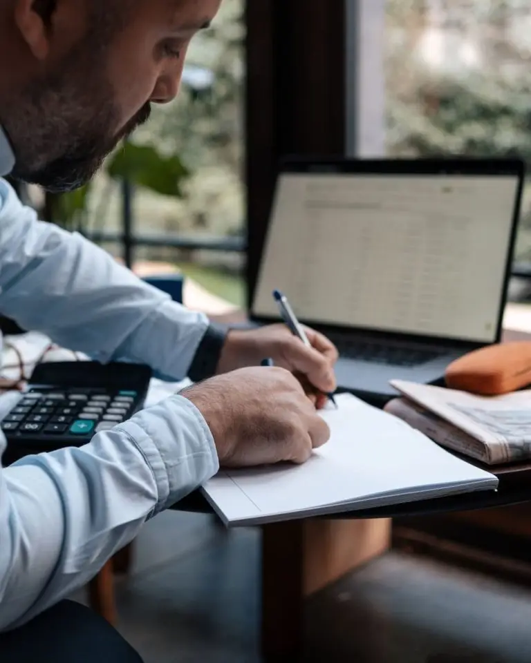 A man writing in a notebook while using a laptop and calculator at a desk.